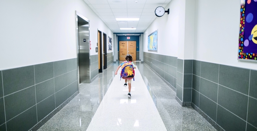 A young kid is wearing a backpack and running down the hallway of their new school.