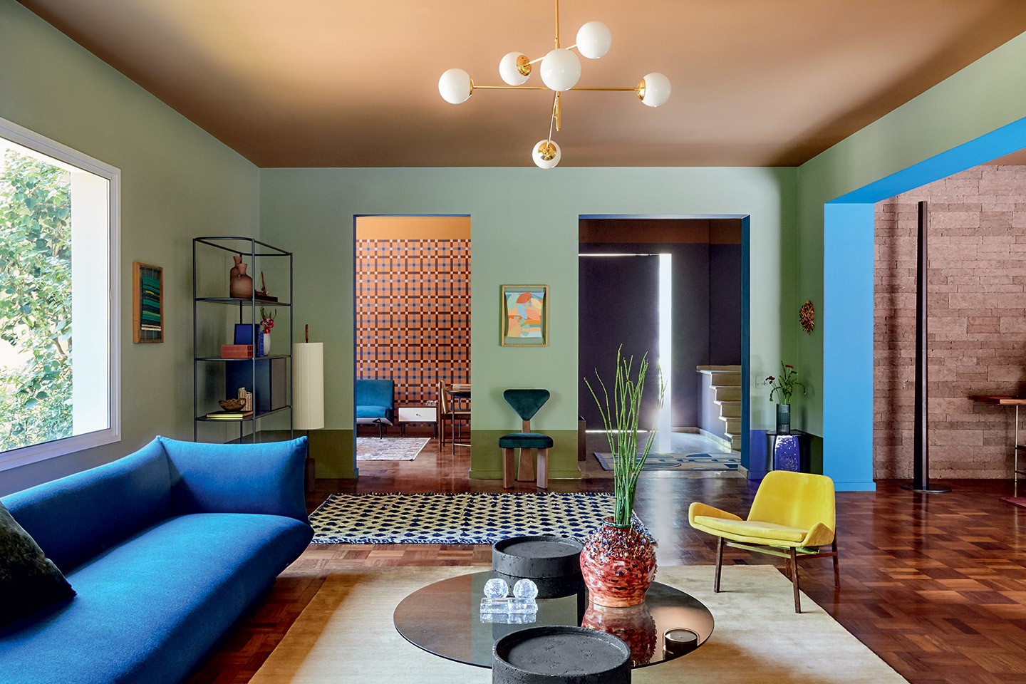 Decoração do Casa Vogue Experience 2021: Sala com piso de taco de madeira, sofá azul, poltrona amarela, mesinha de centro de espelho redonda, paredes verde menta, luminária pendente com design moderno e objetos modernos decorativos.