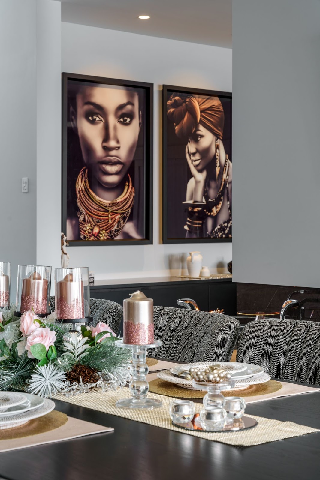 Stylish dining area decorated for a festive occasion, with gray upholstered chairs, a centerpiece of candles and flowers, and large framed portraits of African women adorning the wall.