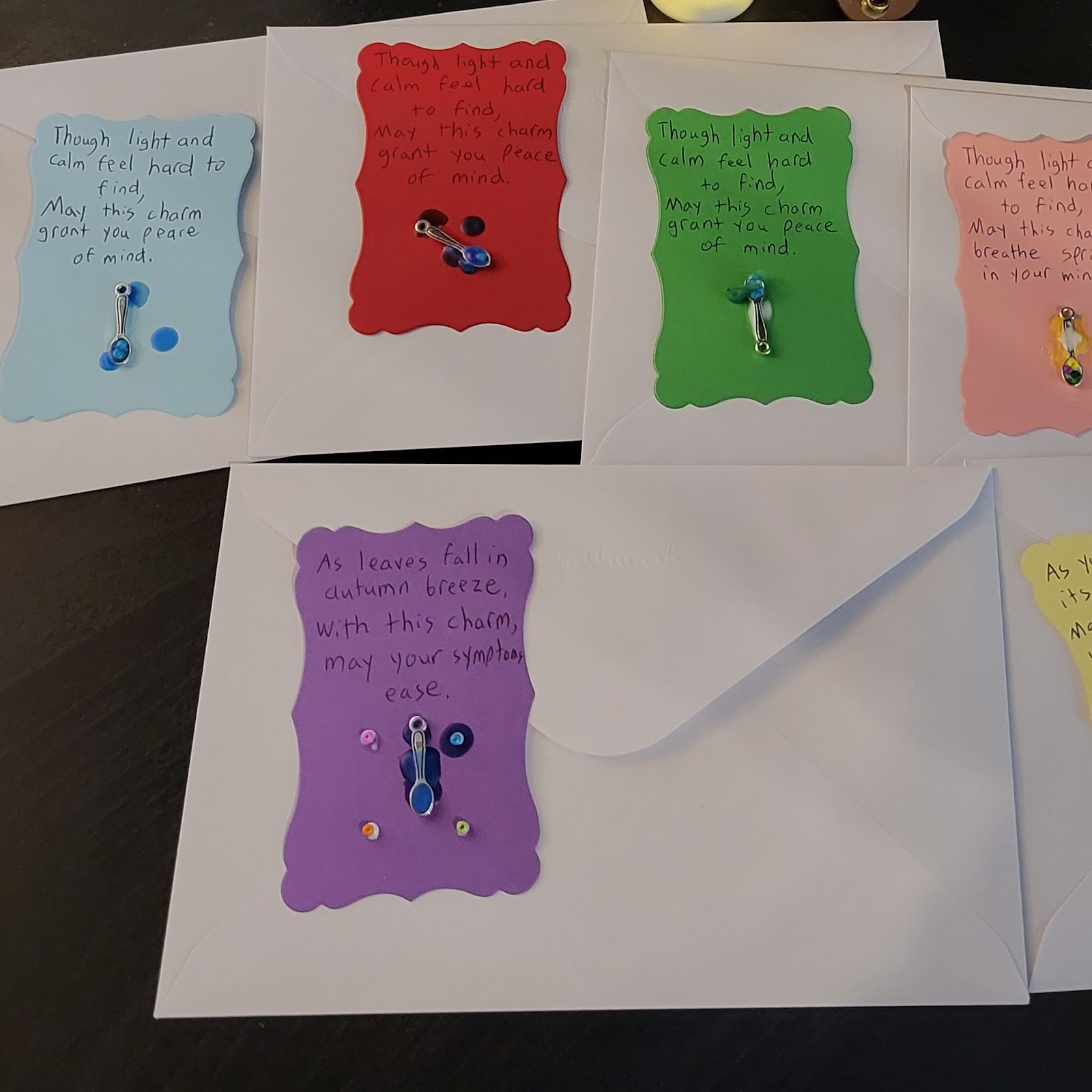 Image description: Envelopes laid out with decorated charms on top as they dry. Each charm includes a colorful piece of cardstock, on which are glued tiny spoons and beads, and each has candle wax dripped on top. Each charm also has an invocation written on it. The various sample invocations are further down in the post.