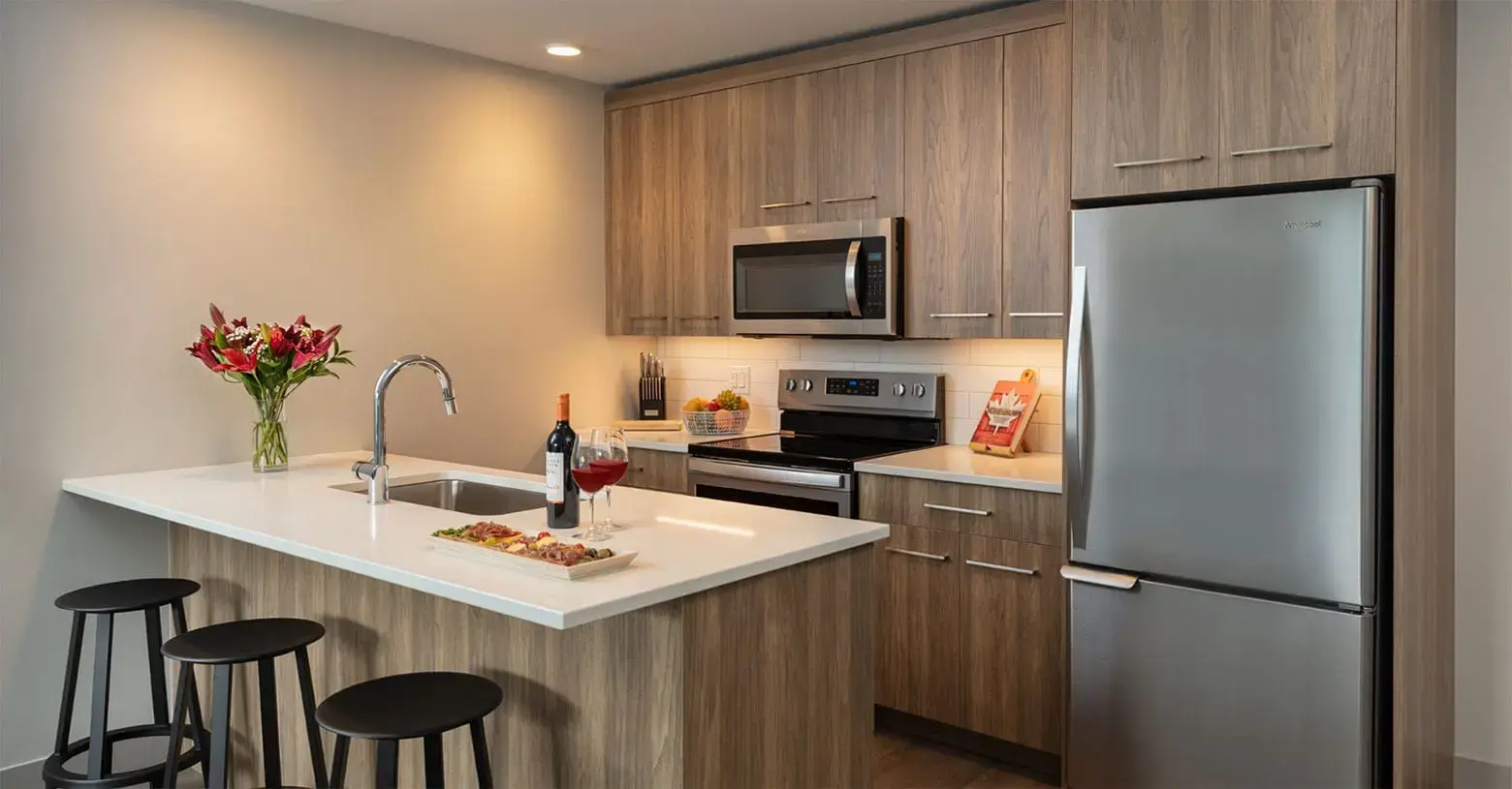 A kitchenette with stainless steel appliances at the Shore Kelowna; on the island countertop are two glasses of wine and a charcuterie board. 
