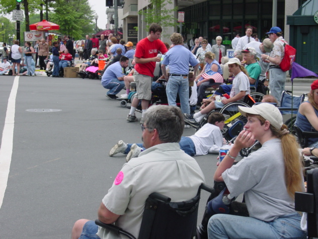 People block a city street, most using wheelchairs, looking into the intersection. 