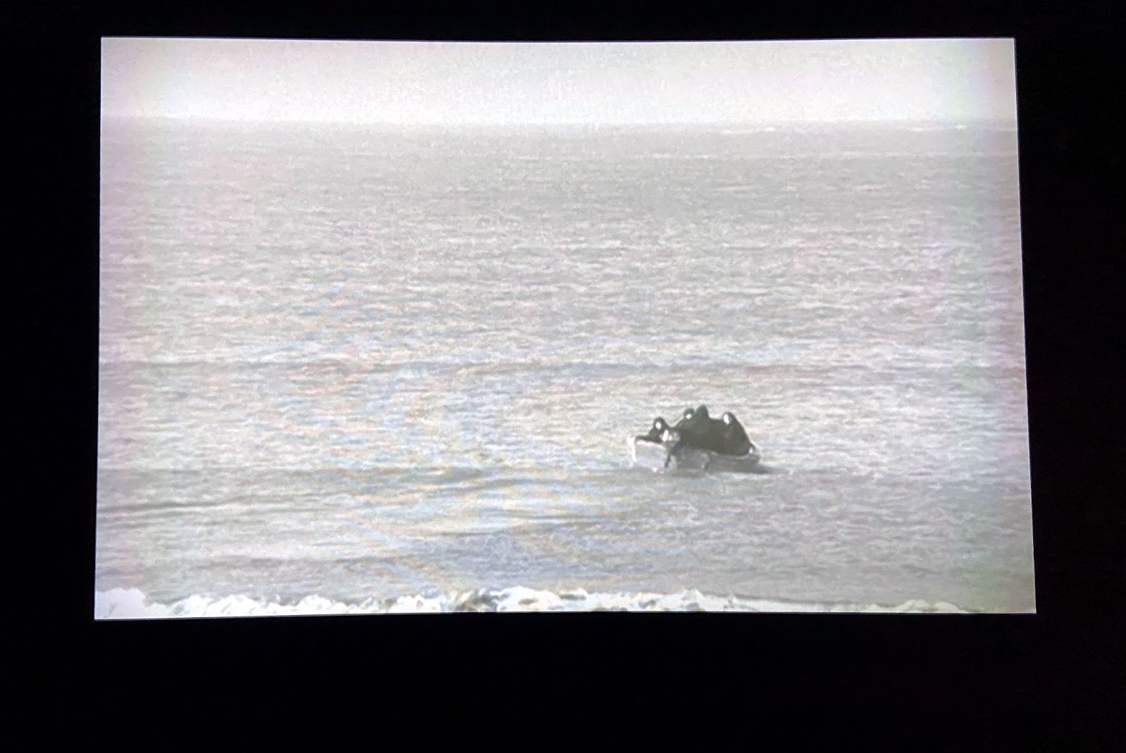 Image: Shirin Neshat, Rapture (still), 1999, two-channel video installation, 16mm black and white film transferred to video. A photograph of a video projection on a black wall. A small motorboat holds at least four women, all wearing black chadors. The boat is on sea or a large body of mostly still water. Small waves crash at the shore at the bottom of the image. Photo by Jessica Hammie.