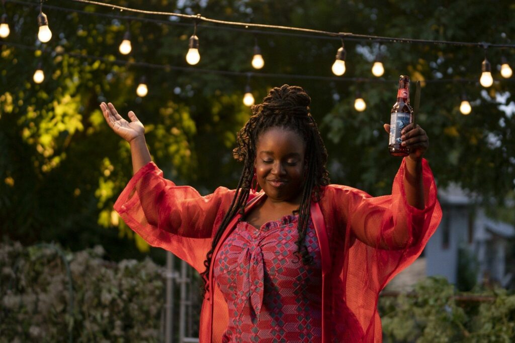 Fran in Shrill. Image: Fran stands in her backyard, wearing an orange and purple print jumpsuit, which ties at the bust. She wears a sheer orange cardigan over her shoulders. Her hair is dark brown and braided corn row style away from her forehead, half up off her shoulders. She stands carefree, holding a beverage in her left hand, shrugging. She is smiling,  eyes closed.