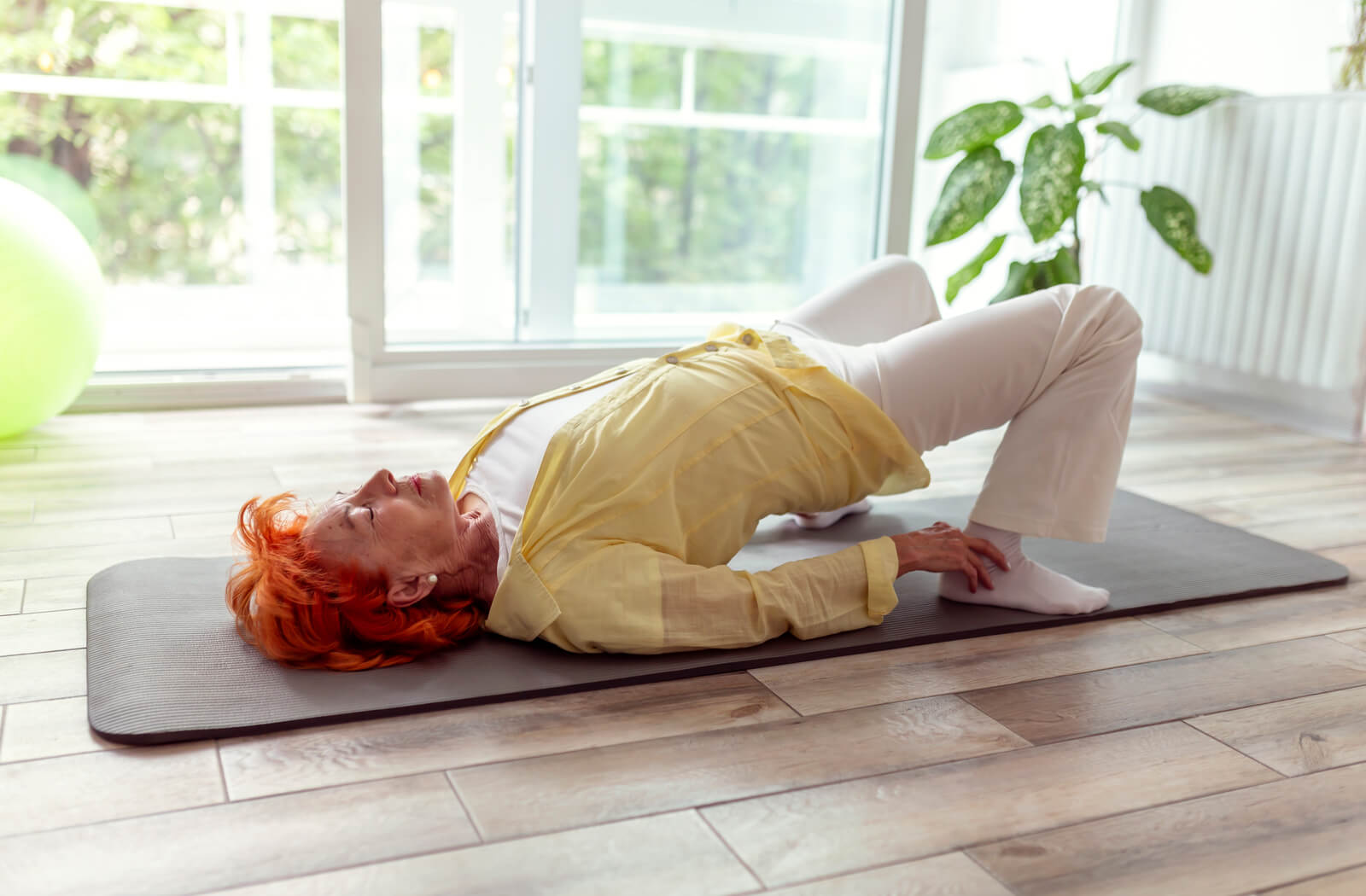 A senior woman lies on a yoga mat, performing a bridge.