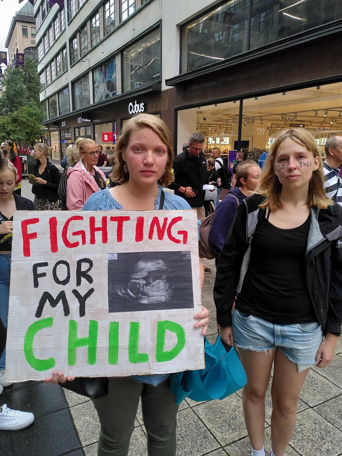 Two mothers look to camera, one bloodshot with tears. She holds a banner with a baby scan that reads 'fighting for my child'