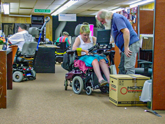 A crowded office environment where a man with white beard stands over a woman in a wheelchair. 