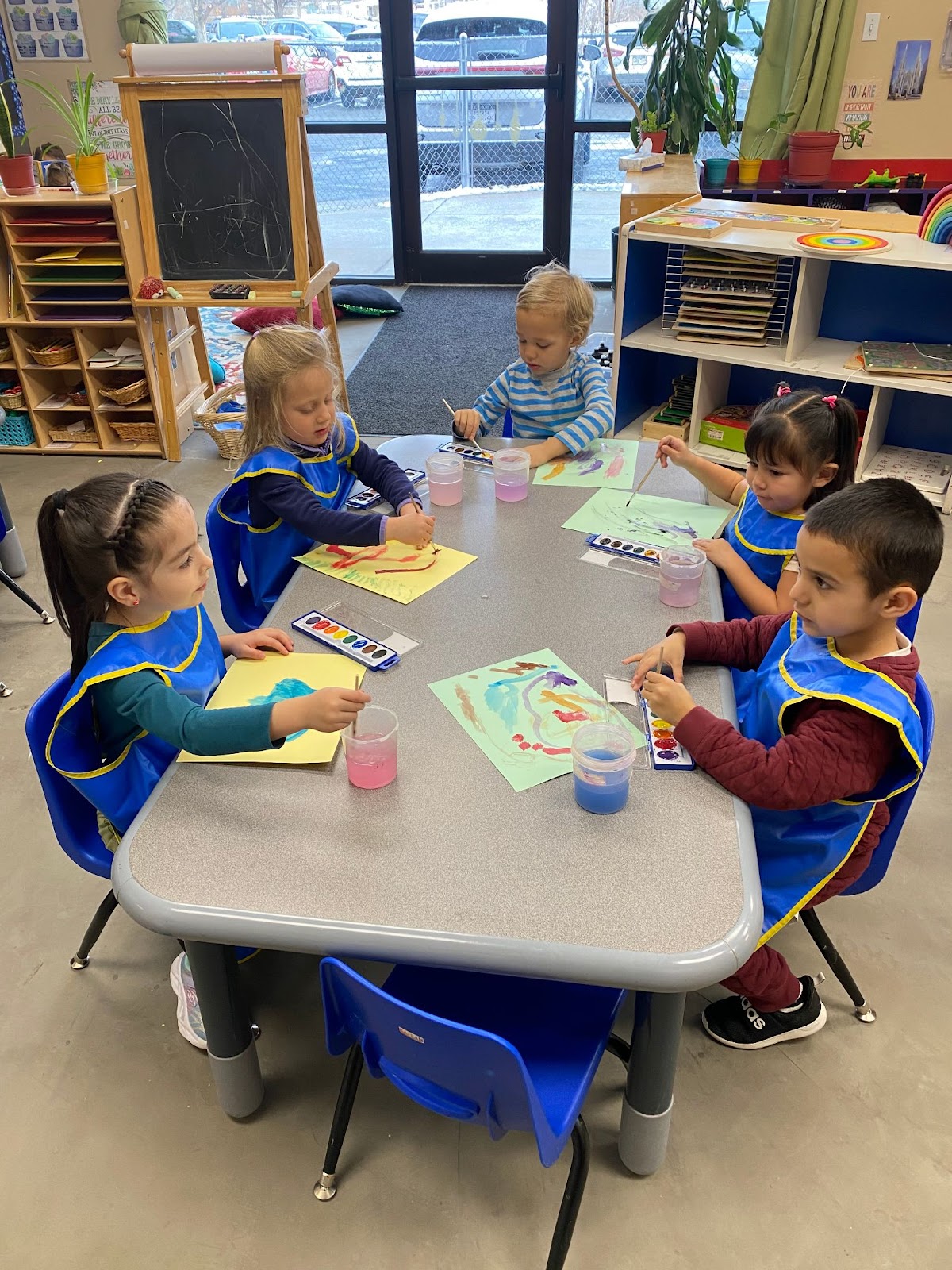 A group of five preschoolers in blue paint smocks sit around a table while watercolor painting
