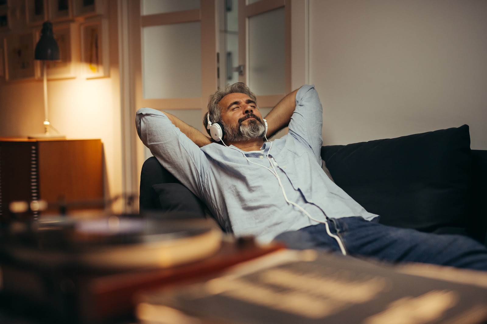 Homem de cabelos e barba grisalhos deitado em um sofá preto, com os braços para trás da cabeça e ouvindo música.