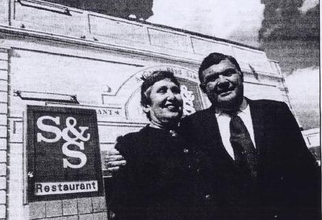 Black and white newspaper phot of a woman and man dressed in dark clothing next to a sign that reads S&S Restaurant