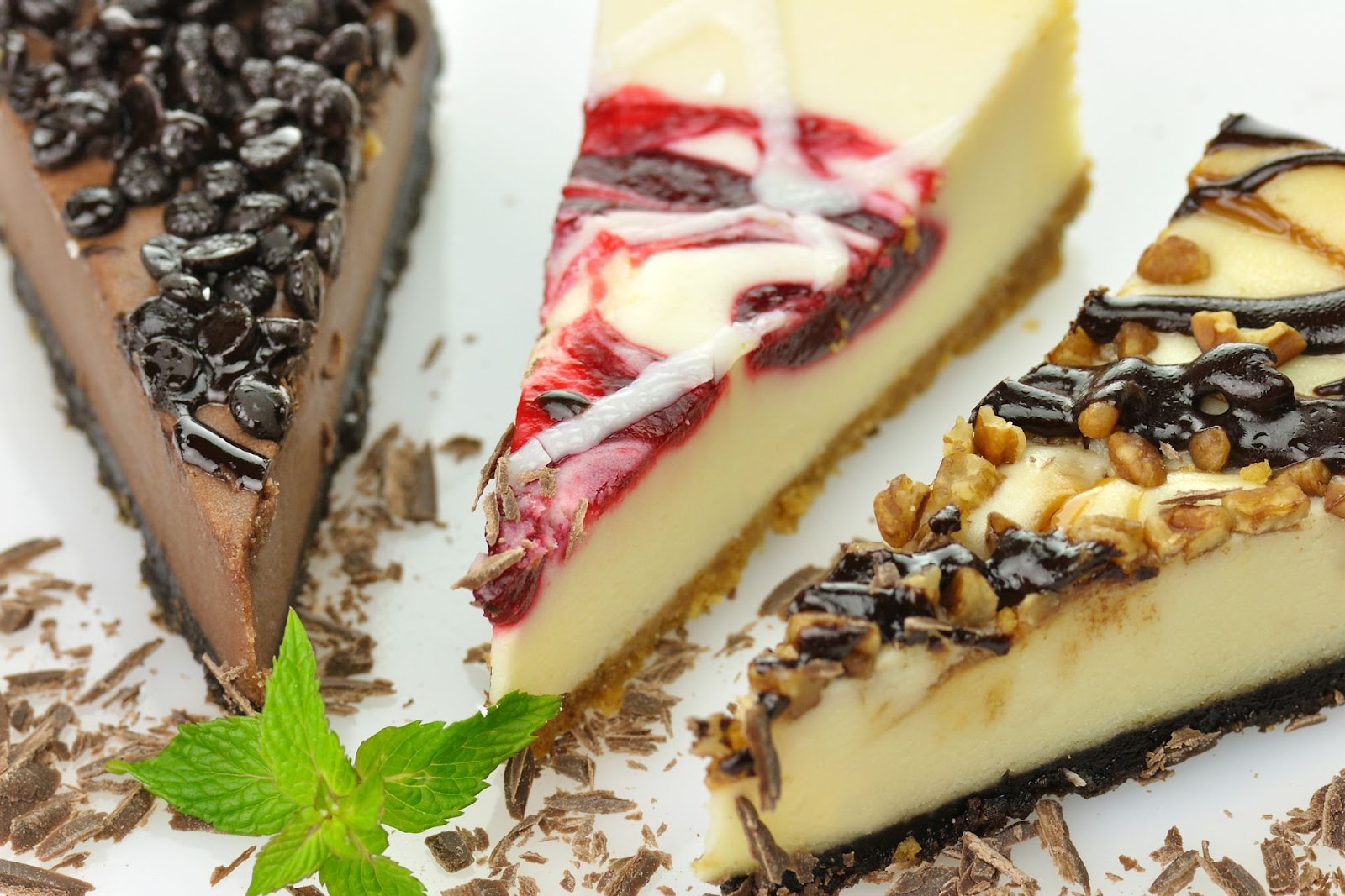 Three slices of cheesecake in a assortment of flavours surrounded by chocolate shavings and a mint leaf as decoration 
