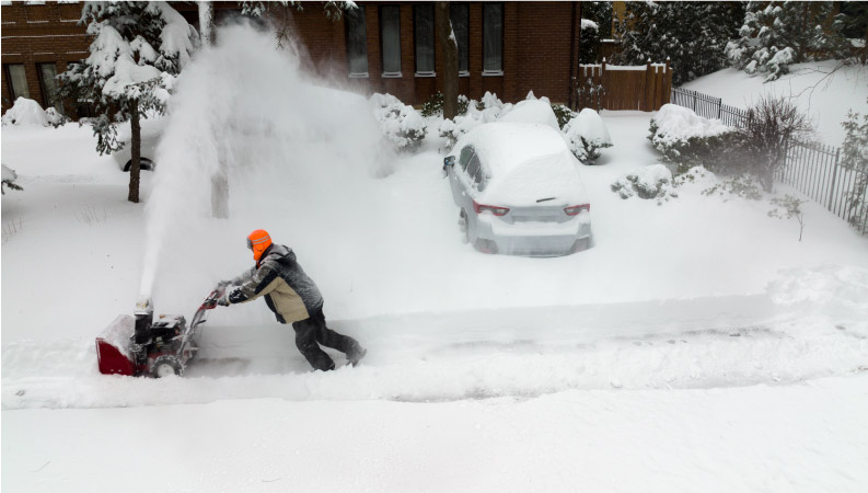 A man, bundled up in winter clothes, is using a snowblower to clear deep snow from in front of his home in Canada.