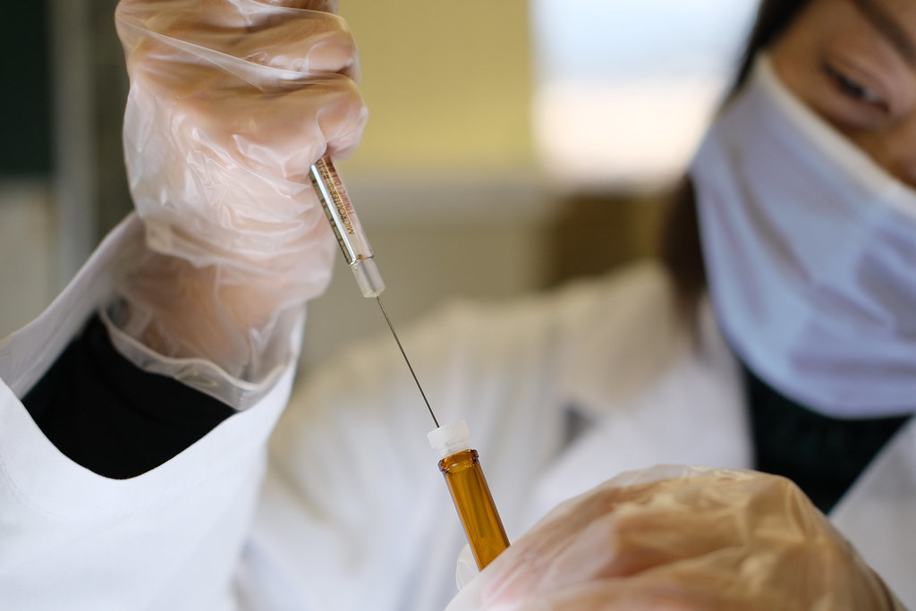 A woman in a lab coat with a face mask on and rubber gloves is using a syringe to withdraw some type of amber colored liquid from a vial.