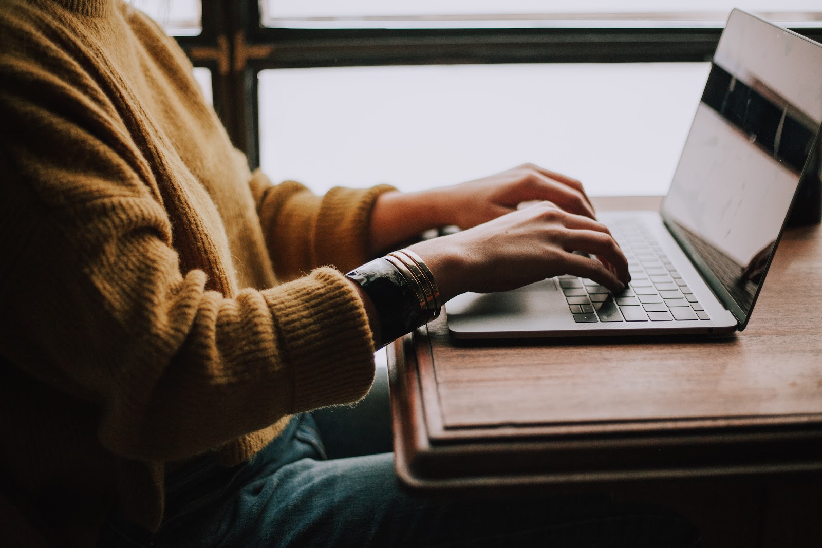 a person with a mustard jumper sits and types on a laptop