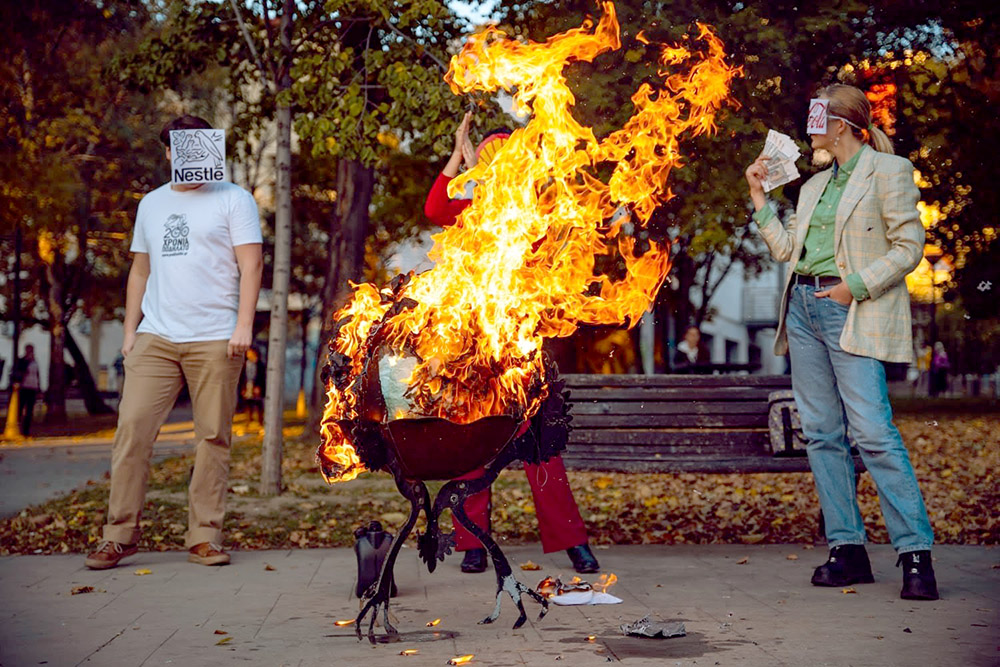 Rebels with corporate logos for masks watch a model Earth burn on a BBQ