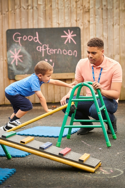 young boy climbs on jungle gym in an outside play area as his male teachers crouches down beside him and watches. In the background, there is a wooden fence with a chalkboard hanging. The message 'good afternoon' is written on the chalkboard.
