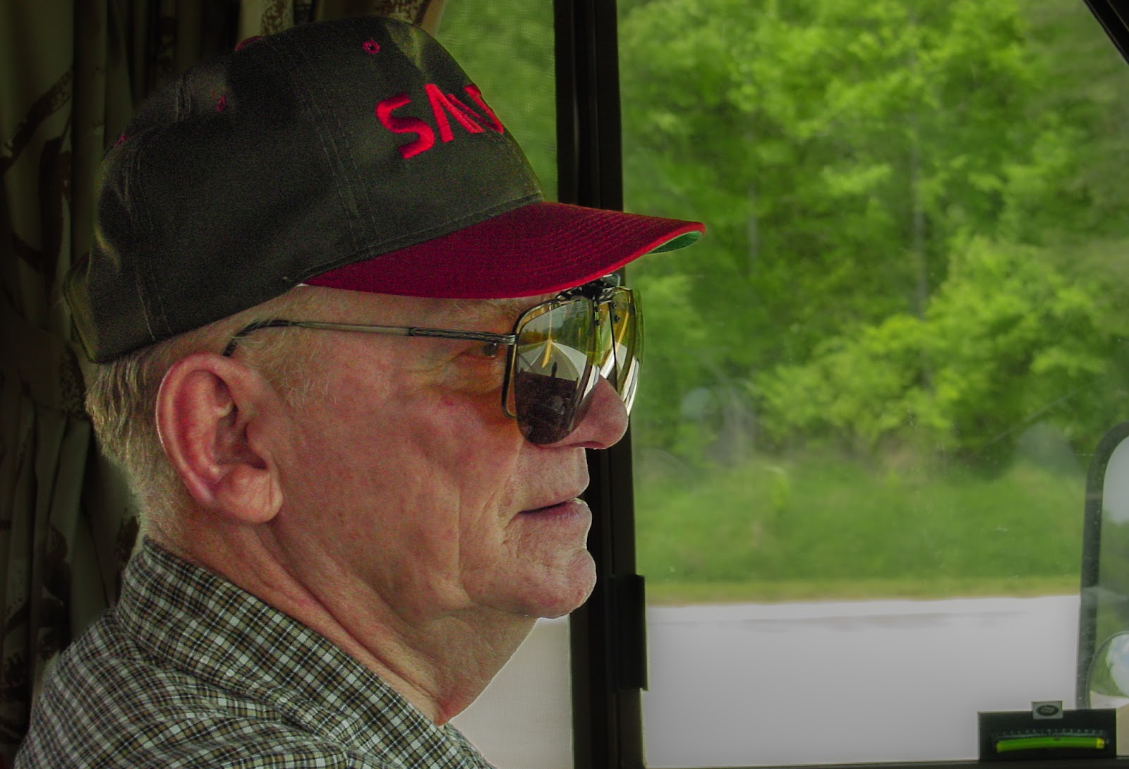 Man wearing cap and glasses, driving a large vehicle. 