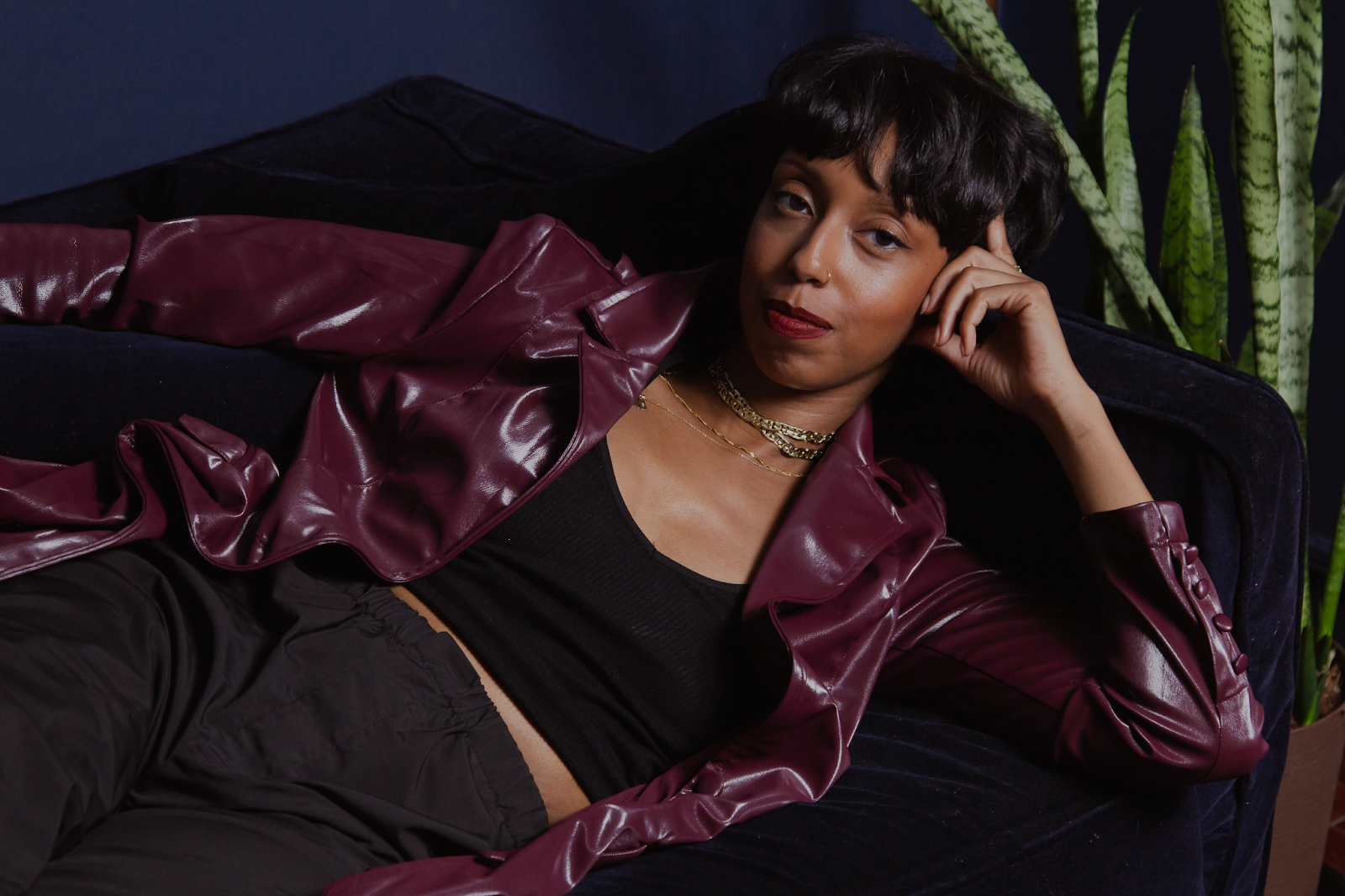 Image: The portrait features Barédu lying down on a dark blue couch in front of a dark blue wall. Behind her, to the right is a snake plant. She has on a burgundy jacket, a black top, and black pants and is wearing gold necklaces, bracelets, and earrings. Image captured and edited by Joshua Johnson.