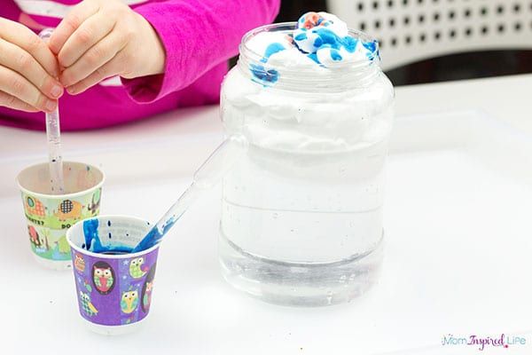 two small paper cups with blue paint-stained water, a large jar filled with shaving cream with blue paint on top, and a child's hands on a dropper in one of the paper cups