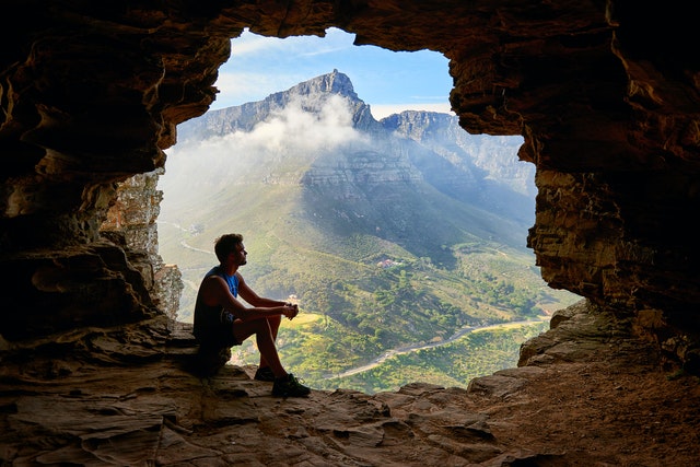 Man seated at the entrance of a cave overlooking a valley below