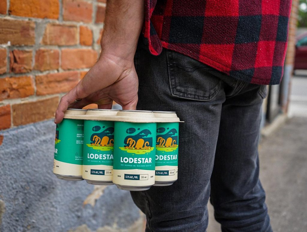 A person standing holding a six pack of Elora's Lodestar beer that's held together with a beige compostable six pack holder.  The person is wearing black jeans and a red and black plaid shirt.