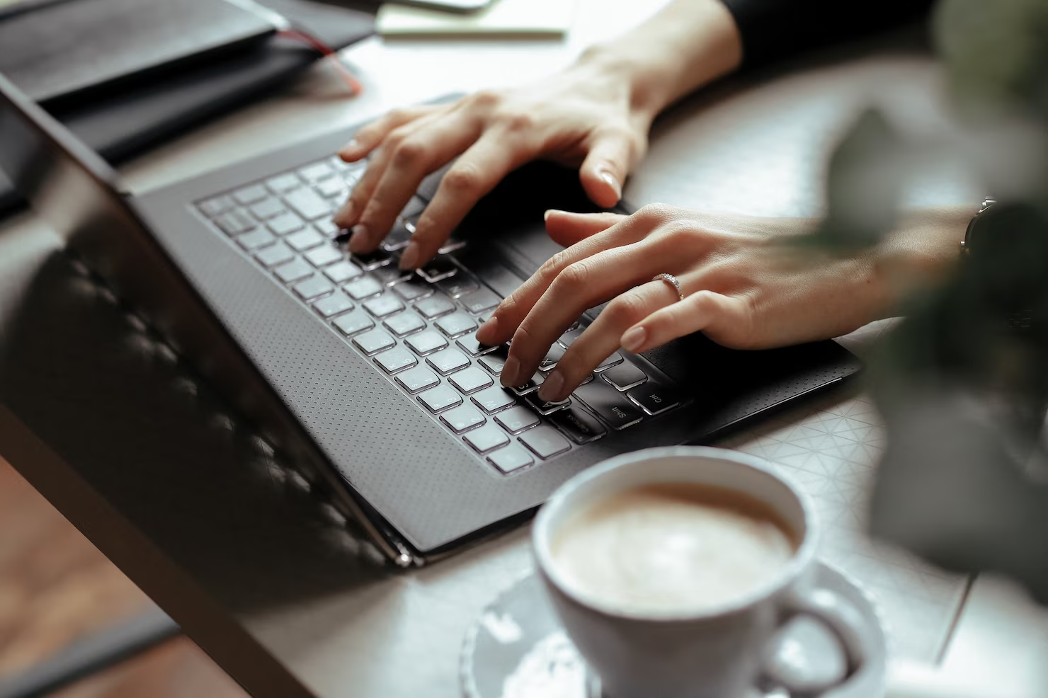 mãos femininas digitam em um notebook preto, acompanhado por um café ao lado em uma xícaras e pires da cor branca
