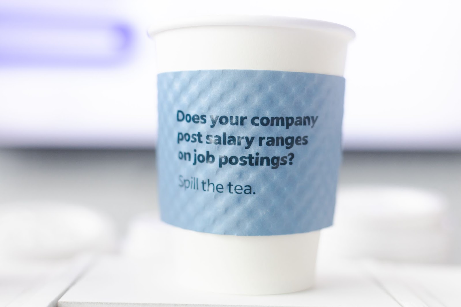Image shows a takeaway coffee cup with a light blue, cardboard sleeve that has the question, "Does your company post salary ranges on job postings? Spill the tea." printed on it.