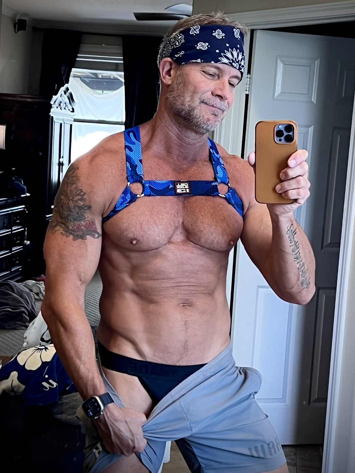 Greg Dixxon wearing a blue camo harness and black jockstrap while pulling down his grey sweatpants for an iPhone mirror selfie