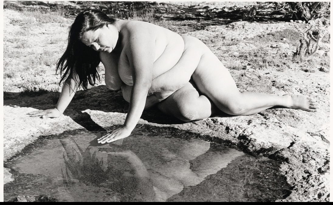 Self-portrait of the artist, nude, kneeling on her hands and knees on a rocky patch of ground.