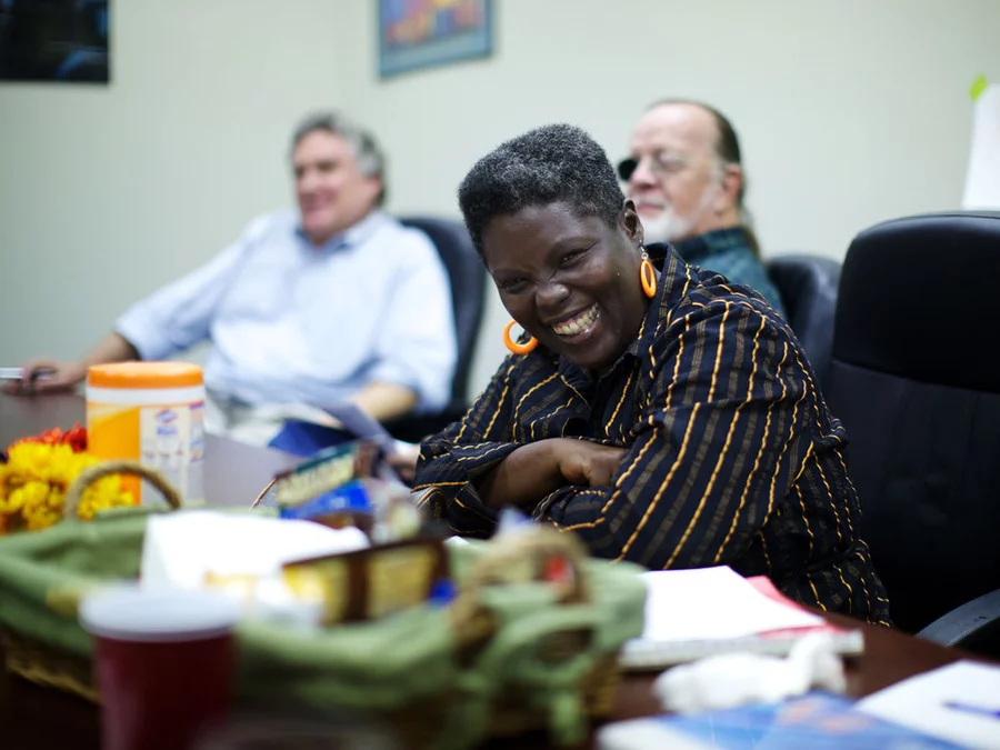 Photo of Lois Curtis, a Black woman with dark skin and short Afro smiling giddily as she sits in a room among friends.