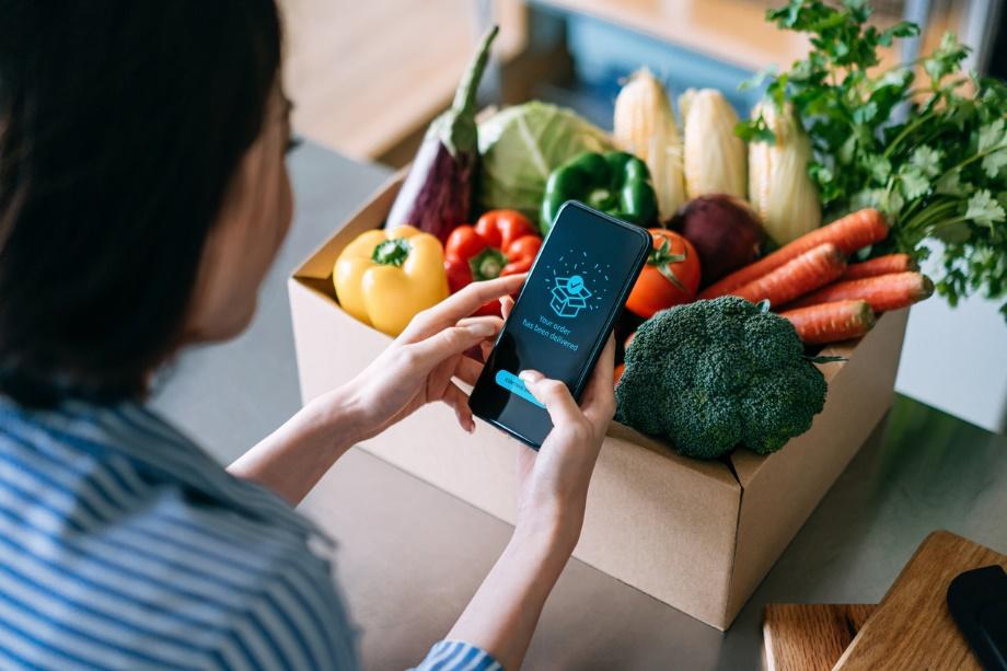 A woman standing over produce she purchased shopping for more sustainable kitchen products on her smartphone.