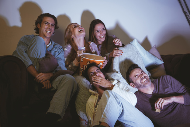 Grupo de amigos sentados no sofá comendo pipoca e rindo de um dos tipos de filmes. 