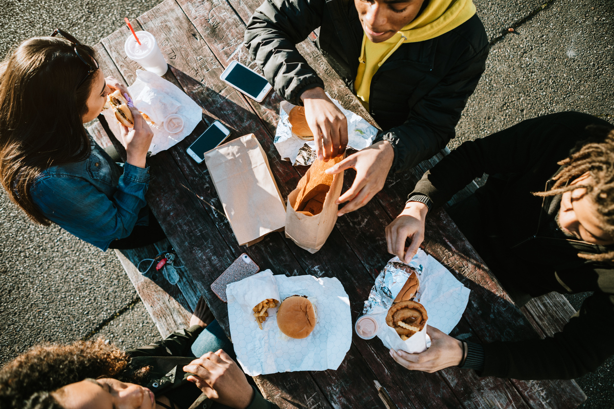 Un gruppo di giovani adulti che mangia al fast food all'aperto: un esempio di abitudini alimentari malsane che possono causare l'obesità.