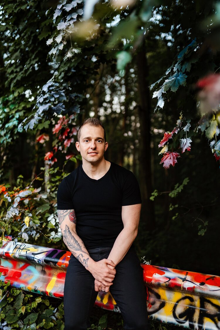 Ryan van Loan is a white masc with close-cropped light brown hair and a large tattoo of a feather on his forearm. In this image, he's leaning against a graffiti'd guardrail in front of a lot of trees.