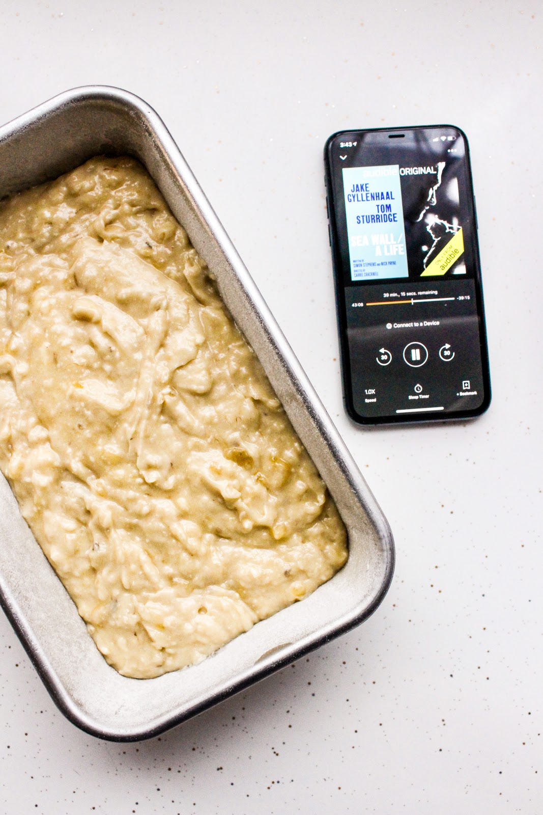 Audio book playing on a phone next to a loaf pan filled with banana bread batter.