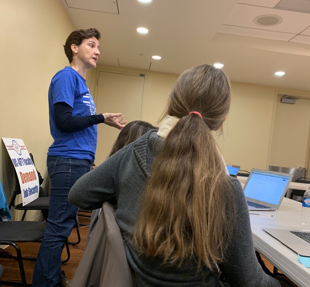 The image shows a woman in a blue UC-AFT t-shirt standing in a conference room and speaking with a serious face. People sitting at tables turn to watch her speak.