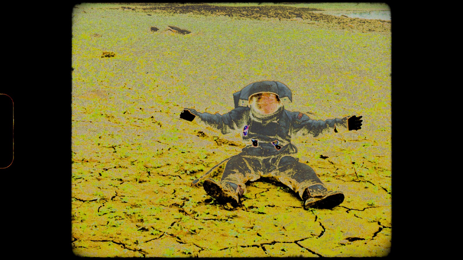 From the film Notes from Eremocene, a person in a astronaut suit sits on the cracked earth, hands outstretched. Yellow particles cover the suit and surrounding environment.