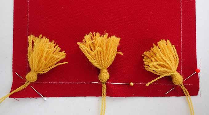Three golden yellow wool tassels, pinned at each corner and at the center or the edge of a square piece of red wool fabric.