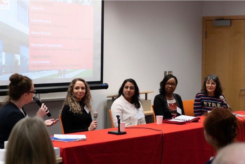 5 people are sitting at a table, 4 of which are panelists. The person from on the left is holding a microphone. 