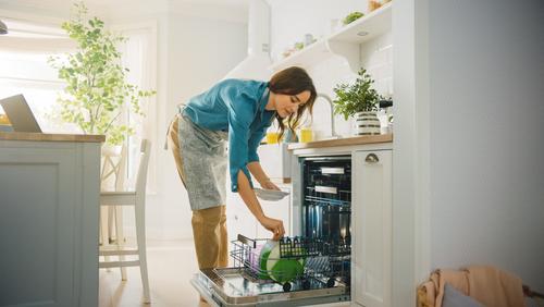 WeServe, a woman bending down to put the dishes in the dishwasher to clean them in a white and wood kitchen