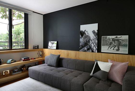 Ambiente com parede de fundo preta, móvel planejado de madeira formando prateleiras pelo ambiente e sofá cinza com almofadas.