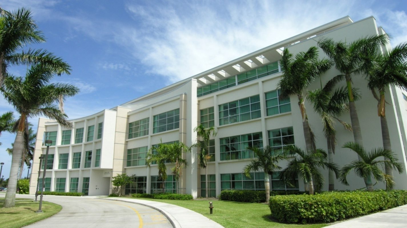 A photo of the Florida Atlantic University Charles E. Schmidt College of Medicine campus, featuring a modern building with glass walls and a courtyard with palm trees.