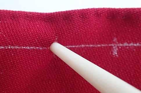 A close up shot of the bone awl pierceing the red wool layer at one of the tick marks indicating a drawstring hole. One of the additional tick marks is visible to the right of the awl. Very faint stitches can be seen on the top edge.