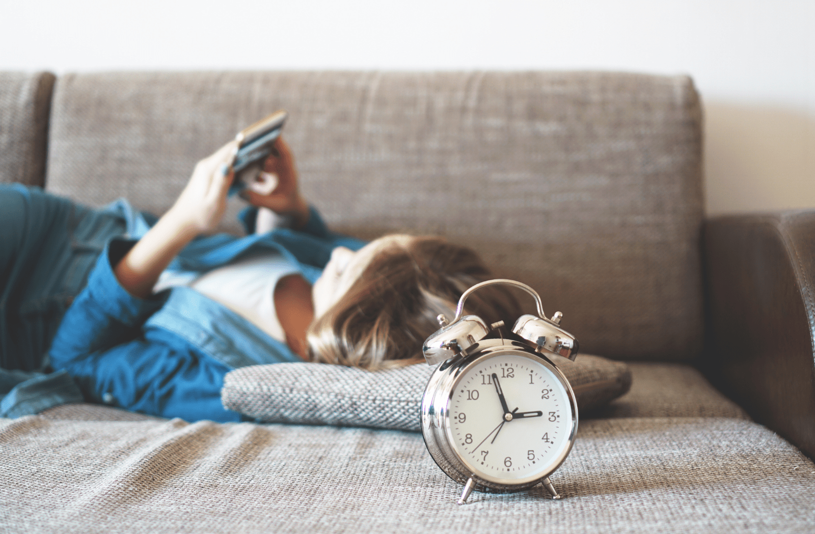 A female teen laying on a couch and looking at her cell phone with a clock nearby.