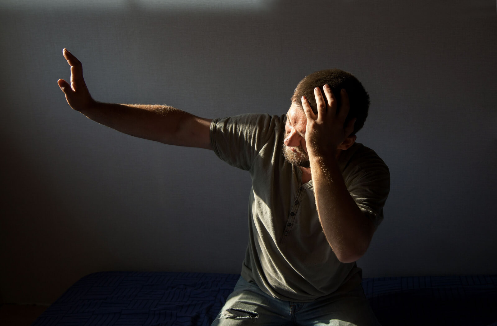 A man sitting on his bed trying to block out sunlight from his window using his hand.