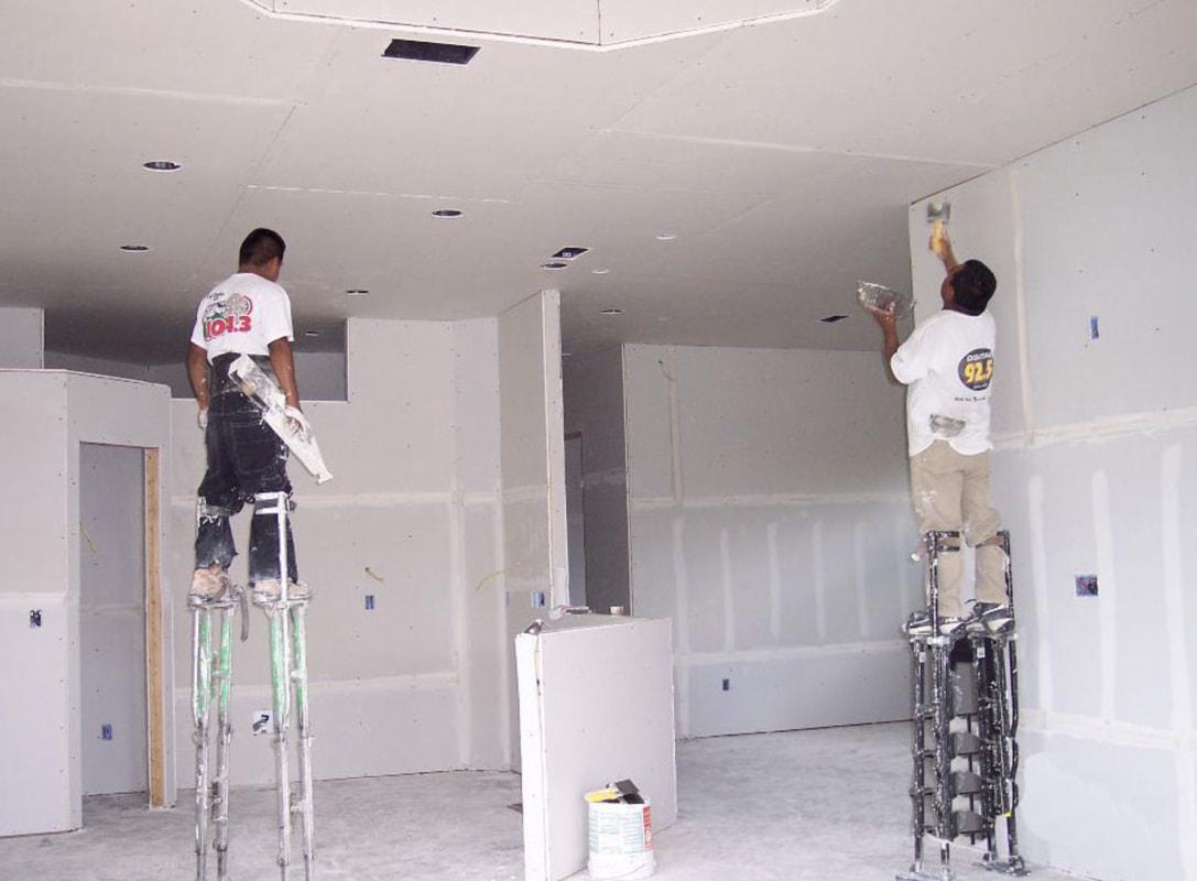 Two contractors standing on stilts working on a drywall.