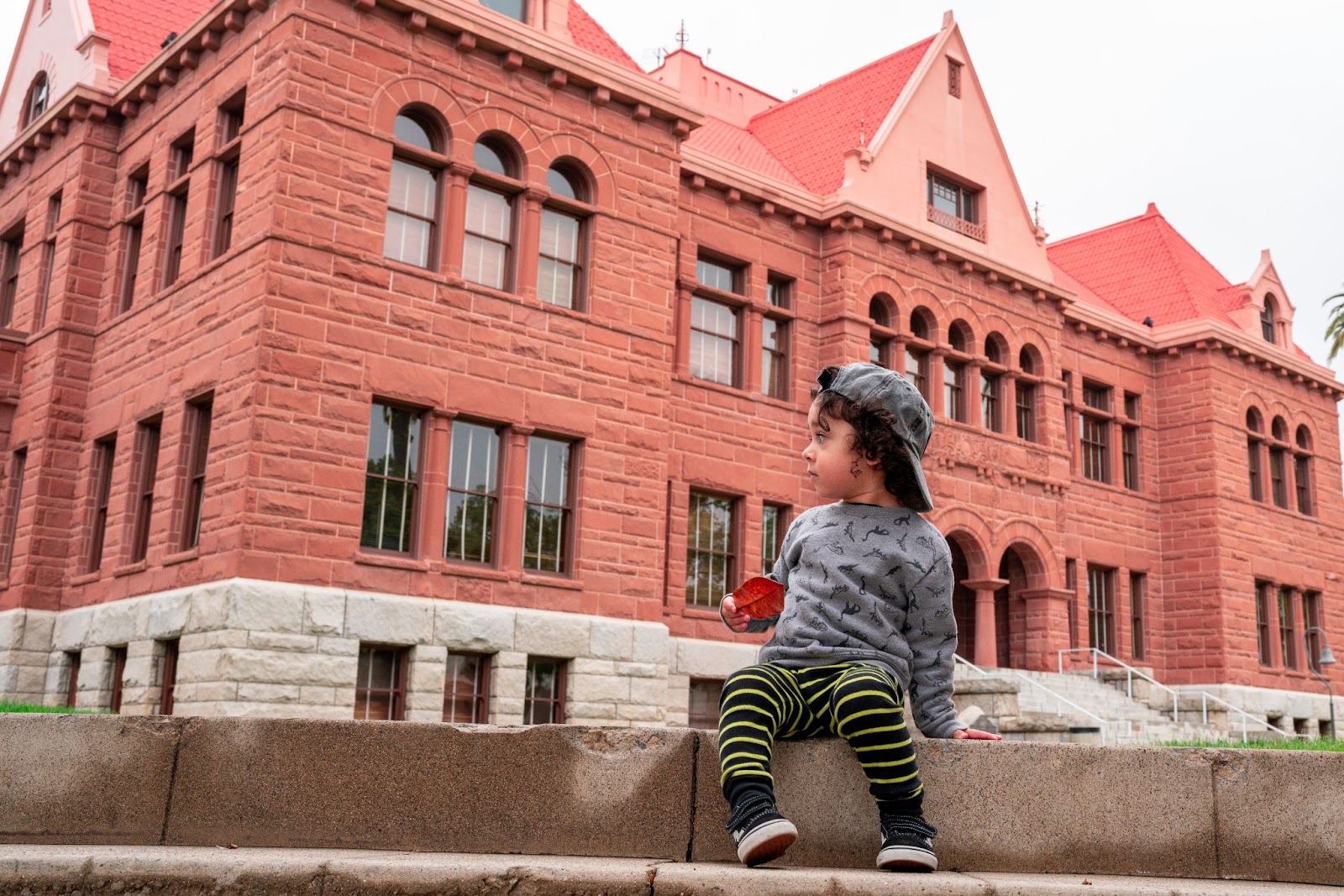 An image of a little boy wearing a long sleeve shirt and leggings and a backwards baseball cap looks off to the left. He is sitting on a ledge in front of the old OC courthouse, a red brick building with a red roof.
