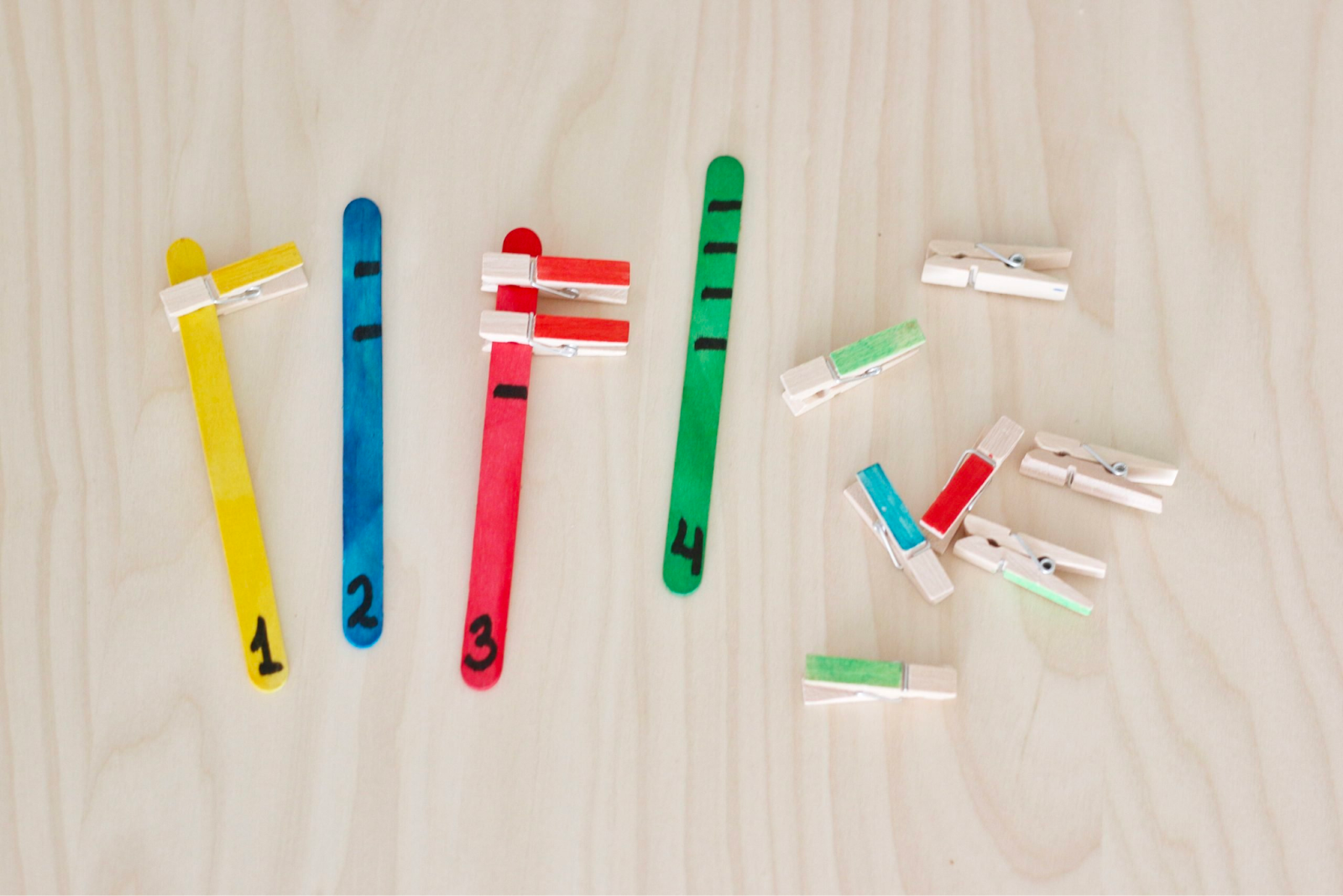 Four popsicle sticks: the first painted yellow, with the number 1 at the bottom; the second painted blue, with the number 2 at the bottom; the third painted red, with the number 3 at the bottom; and the fourth painted green, with the number 4 at the bottom. There are seven clothespins to the right of the popsicle sticks. 