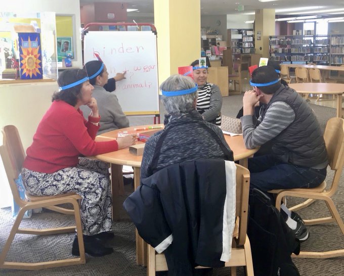 Adults playing Hedbanz, pointing at a whiteboard with the words: binder, wear, animal