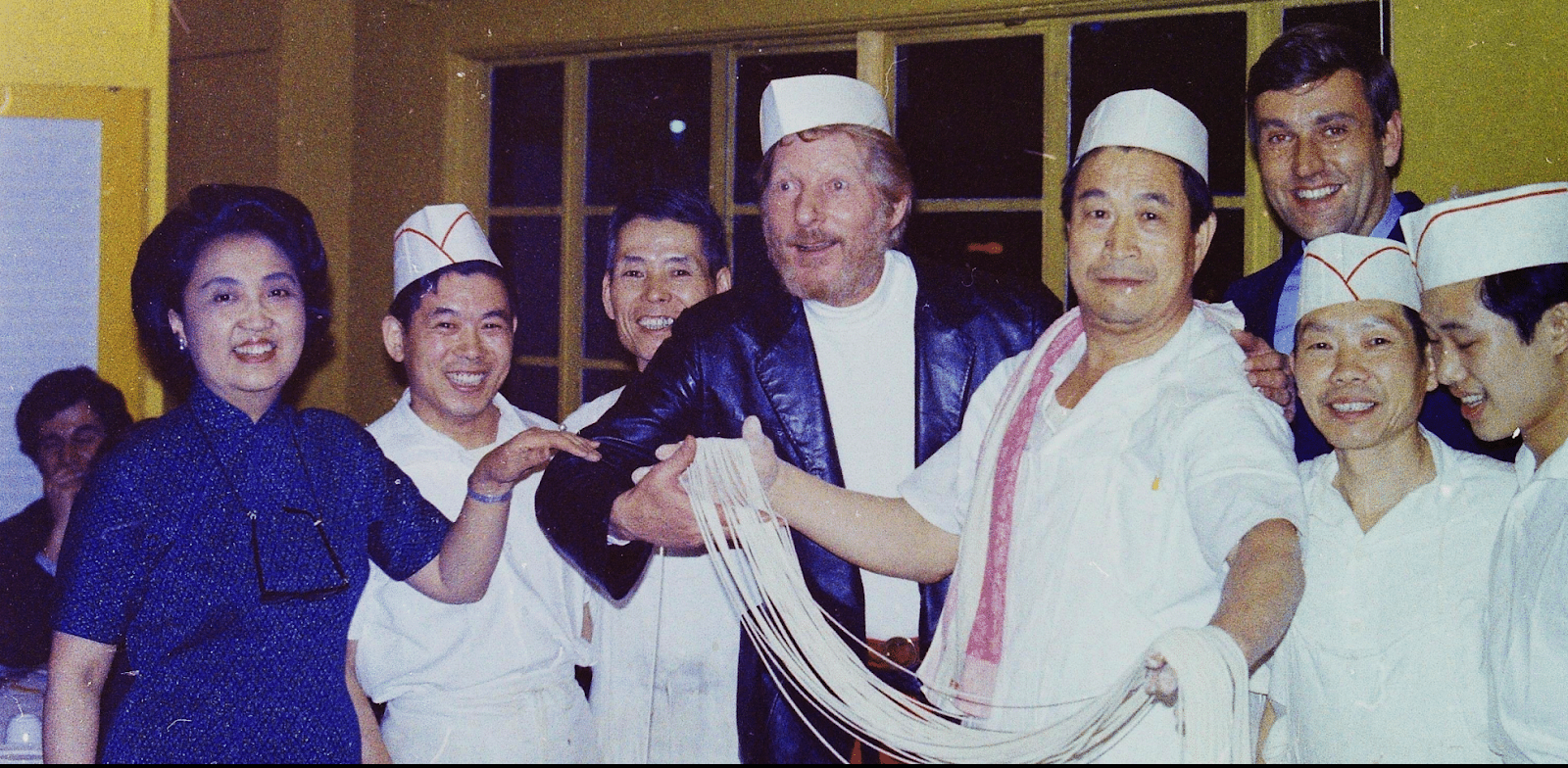 Group of people smiling. Woman at left with dark hair in a blue dress has glasses hanging from her neck. Man in center with dark jacket is helping hold a long stretch of noodles held by man in white shirt and hat.